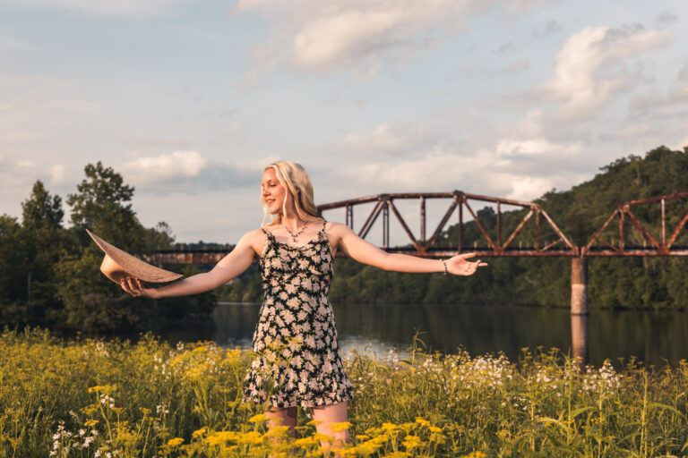 A Hardin valley high school high school senior poses in a field of flowers in front of a train trestle that goes over a river